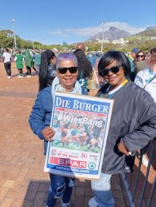 Two women smiling and holding a Die Burger rugby poster with the hashtag WiesBang during a DBD Travel group outing