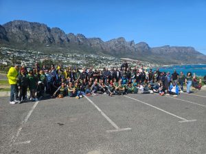 A large group of DBD Travel tourists posing together in a coastal parking area with the iconic Twelve Apostles mountain range and blue ocean in the background.