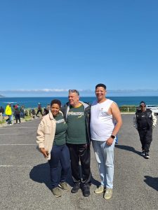 Three smiling travellers wearing South African Springboks gear standing together at a scenic coastal lookout point during a DBD Travel group tour.