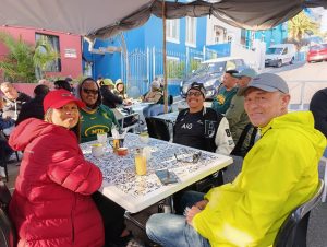 A group of travellers wearing South African Springboks and New Zealand All Blacks supporters gear, enjoying a meal together at an outdoor café on a sunny day.